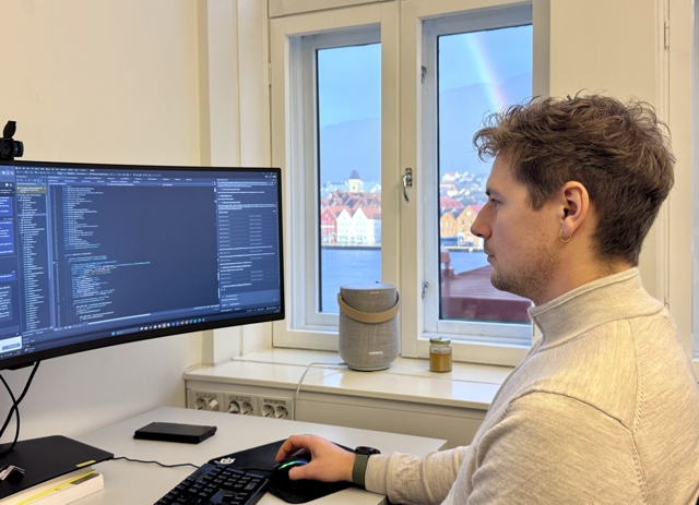 Sturle koder ved skrivebordet sitt i Bergen med bryggen som backdrop honning i vinduet og en regnbue i bakgrunnen